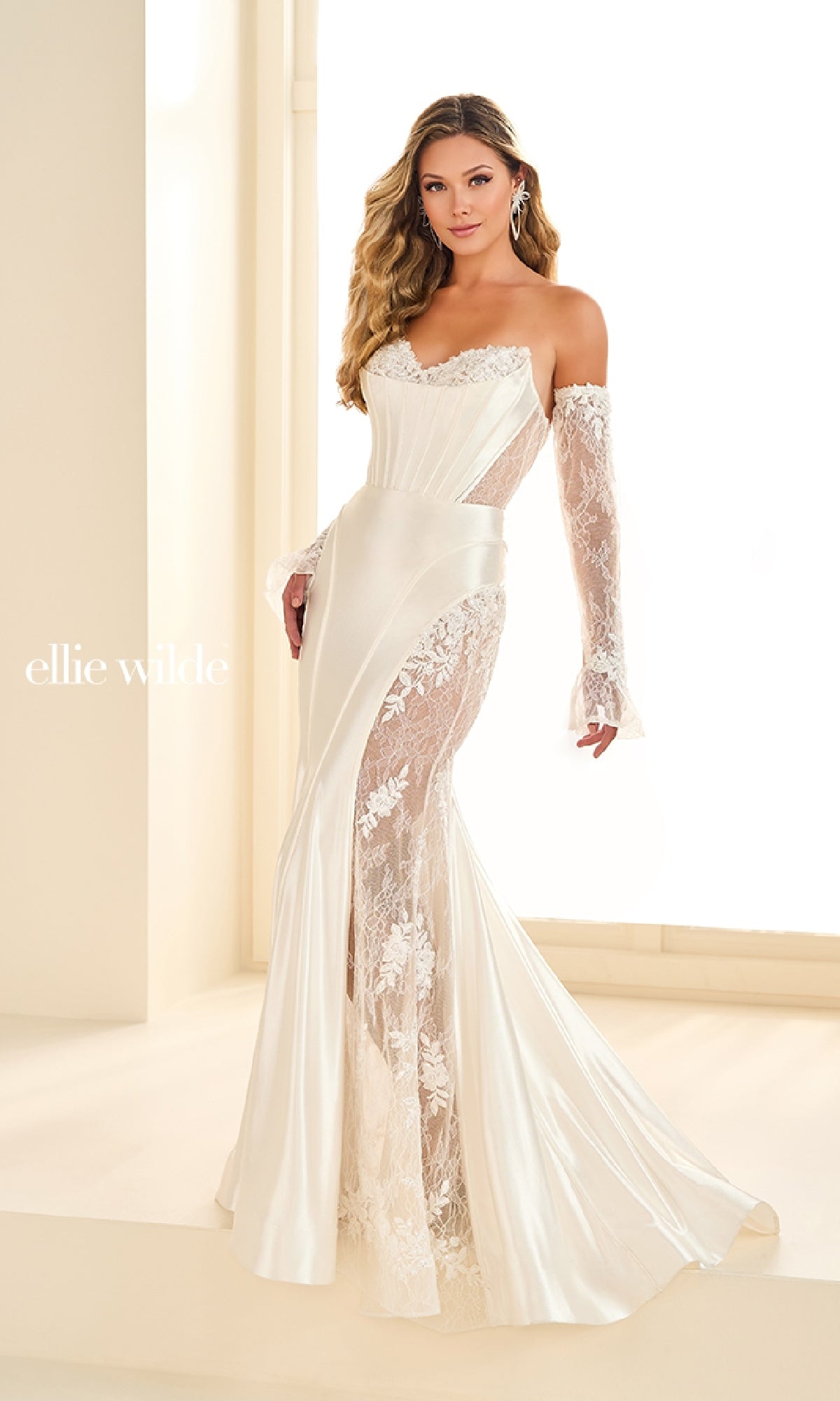 A woman poses indoors wearing the Ellie Wilde EW36010—a long, off-the-shoulder white mermaid prom dress featuring lace sleeves, lace panel details, and a flattering corset bodice.